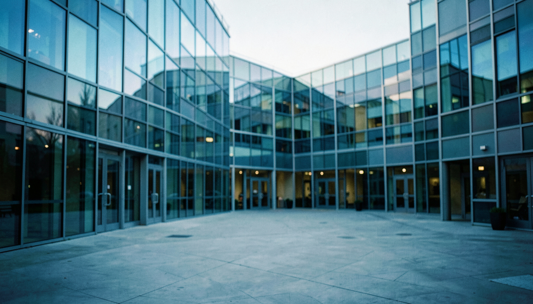 A wide-angle view of a modern research campus courtyard with glass facades reflecting soft morning light, muted blues and grays